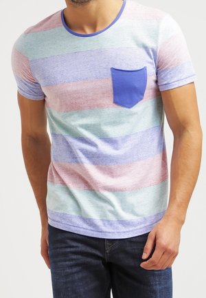 Mann, der ein pastellfarben gestreiftes Kurzarm-T-Shirt mit einer blauen Brusttasche und dunkelblauen Jeans trägt, steht mit einer Hand an der Seite.