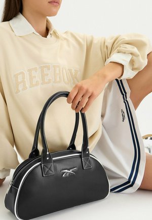 Kobieta w beżowej bluzie Reebok i białych szortach trzyma czarną torebkę Reebok z białymi lamówkami i logo.