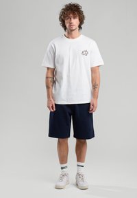 Valkoinen pyöreäkauluksinen t-paita, jossa on pieni musta grafiikka rinnassa, yhdistettynä tummansinisiin shortsihousuihin ja valkoisiin lenkkikenkiin, joissa on vihreitä yksityiskohtia.