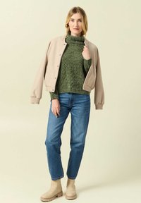 Beige bomberjacka med tryckknappar, grön stickad polotröja, blå raka jeans och beige ankle boots. Enkelt design, texturerad tyg.