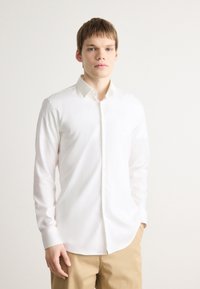 Weißes Langarmhemd mit button-down Kragen, glattem Stoff, slim fit und einem dezenten Markenlogo auf der Brusttasche, kombiniert mit beigen Hosen.