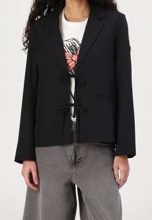 Femme portant un blazer noir avec deux liens sur le devant, par-dessus un tee-shirt blanc avec motif et un jean gris taille haute.