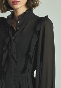 Vestido negro con un cuello y mangas con volantes, adornado con botones decorativos, que presenta una textura de tela ligera y transparente.