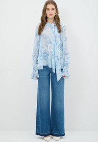 Lyseblå blomsterbluse med løse ærmer og slipskrave, parret med vidde denimjeans prydet med små perler og hvide hæle.