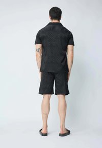 Ensemble composé d'une chemise noire à manches courtes et d'un short, arborant un motif de vagues texturé. Le matériau est léger avec une coupe décontractée, accompagné de sandales à enfiler.