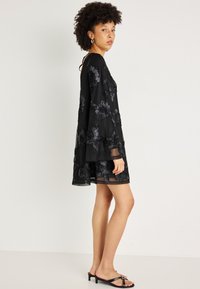 Melnas kleitas ar caurspīdīgām, zvana formas piedurknēm, kurās ir ziedu izšuvumi, slāņveida dizains un paplašināta silueta. Valkāta ar melniem sandaliem.