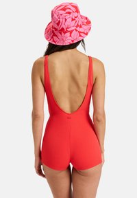 Roter, strukturiert Badeanzug mit tiefem Rückenausschnitt und figurbetontem Schnitt, kombiniert mit einem rosa, blumigen Bucket Hat mit breiter Krempe und lebhaftem Muster.