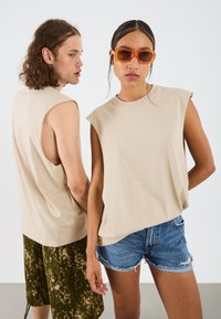 Beige ujjatlan felsők két modellen; az egyik zöld terepmintás sortban, a másik farmer sortban. Kiegészítők között narancsszínű napszemüveg található.