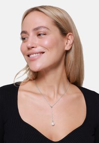 Sølvkjede med flat kjede som har en hengende pendant med en enkelt perle. Modellen har på seg en svart ribbestrikket V-hals topp.