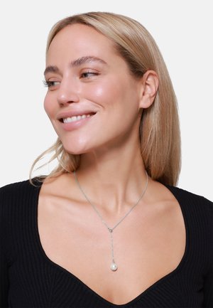Silberne Halskette mit einer flachen Kette, die einen Abgehänge-Anhänger mit einer einzelnen Perle trägt. Das Model trägt ein schwarzes, geripptes V-Ausschnitt-Oberteil.