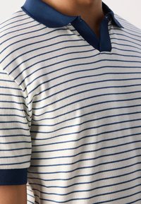 Gros plan sur une chemise blanche à manches courtes côtelée avec de fines rayures horizontales bleu marine, un col bleu marine et des poignets de manches bleu marine.