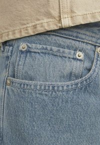 Ljusblå denimjeans med en synlig bakficka, accentuerad av metallnitar och distinkt sömnad. Tyget har en strukturerad yta.