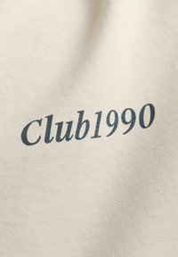 Beige tričko s potiskem "Club1990" v tmavě modrém serifovém písmu. Měkká, hladká textura s klasickým střihem a minimalistickým designem.