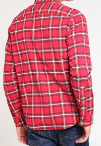 Camicia a maniche lunghe a quadri rossi con strisce nere e gialle, colletto abbottonato e polsini abbottonati. Il tessuto sembra morbido e leggero.