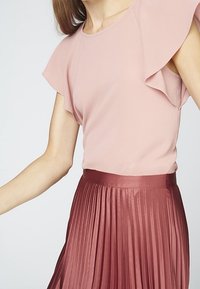 Donna che indossa una blusa a maniche a volant di un tenue rosa chiaro, infilata in una gonna in satin plissettata a vita alta di un profondo colore rosa.