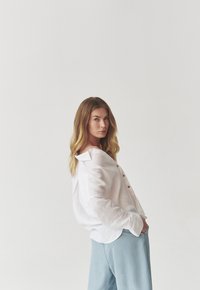 White linen button-up shirt with a collar, featuring wooden buttons and loose fit, paired with light blue wide-leg trousers.