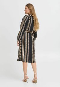 Rochie neagră cu modele verticale în crem și aur. Are o curea, mâneci lungi și o lungime până la jumătatea gambei. Purțită cu sandale cu barete.