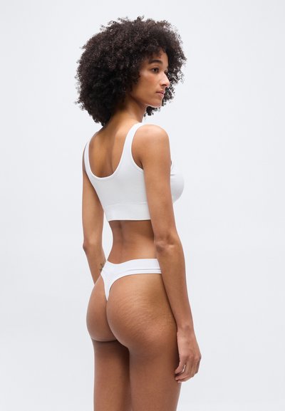 Γυναίκα με σγουρά μαλλιά που φοράει λευκό τοπ και εσώρουχο, στέκεται σε τριών τετάρτων πόζα με την πλάτη, μπροστά σε απλό φωτεινό φόντο.