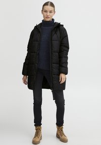Svart pufferjakke med hette, glidelåslukking, quiltet design, båret over en marineblå genser, kombinert med svarte bukser og brune snørebåndsboots.