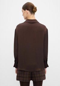 Blusa marrón de manga larga con un corte relajado, textura suave, cuello puntiagudo y puños abotonados, combinada con shorts a cuadros.