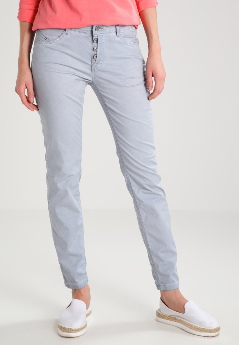 Femme portant un pantalon slim gris clair à fermeture quatre boutons, un top corail à manches longues et des chaussures blanches slip-on, debout sur un fond uni.
