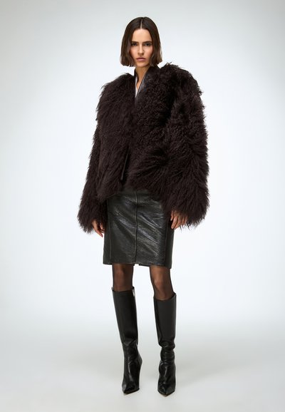 Kobieta ubrana w ciemną, oversize'ową puszystą kurtkę, czarną skórzaną spódnicę, przezroczyste czarne rajstopy i wysokie czarne skórzane kozaki, stojąca na białym tle.