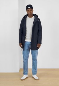 Abrigo azul marino para hombre con capucha, sudadera gris, jeans de mezclilla de color azul claro y zapatillas blancas, de pie sobre un suelo de madera frente a un fondo plano.