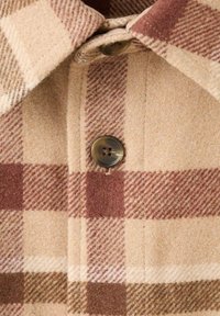 Tela de cuadros beige y marrón con textura suave, que presenta un cierre de botones y detalles de cuello, confeccionada en lana o material similar.