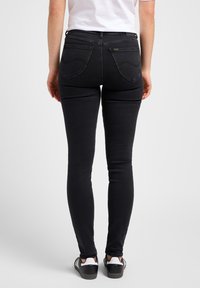 Jean skinny noir avec une taille mi-haute, un design à cinq poches et des détails de couture subtils sur les poches arrière. Le tissu semble lisse et extensible.