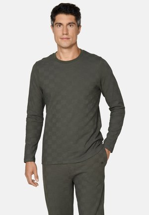 Man draagt een donkergroen shirt met lange mouwen en bijpassende broek met een subtiel geruit patroon, staat met één hand in de zak tegen een witte achtergrond.
