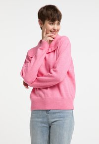 Rózsaszín kötött pulóver laza fit-tel, behajtott gallérral és bordázott szegéllyel. A modellen világoskék farmernadrág van. Puha textúra, laza dizájn.