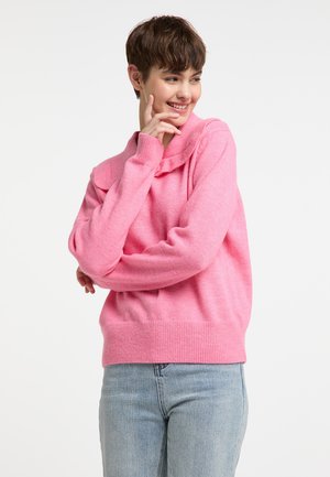 Růžový pletený svetr s volným střihem, s sklopeným límcem a žebrovaným lemem. Model má na sobě světle modré džíny. Měkká textura, pohodlný design.