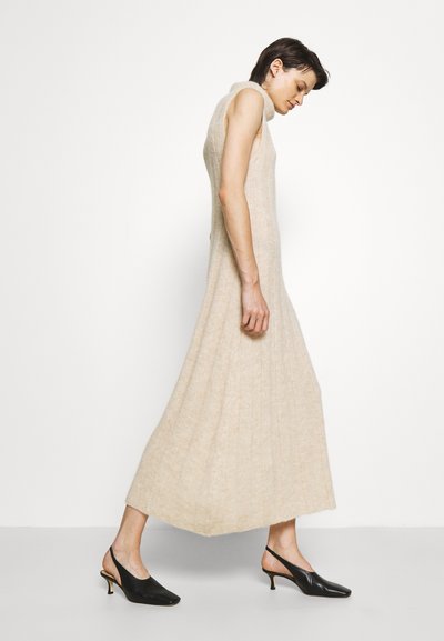 Vestido sem mangas até ao joelho em tricot bege claro com riscas verticais; usado com sapatos de salto alto pretos com abertura atrás.