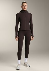 Brun sportjacka och tights med åtsittande design. Jackan har dragkedja och huva. Modellen bär ljusa sneakers och vita strumpor.