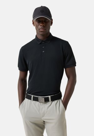 Mężczyzna w czarnej koszulce polo i czapce, beżowych spodniach i czarnym pasku, stojący z rękami w kieszeniach na białym tle.