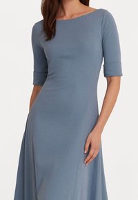 Sininen polveen ulottuva mekko, jossa on venepääntie, kolmannes hihoista ja sileä pinta. Ominaisuuksina käärityt hihojen suut ja levenevä hame.