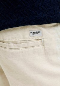 Gros plan sur une poche de pantalon en tissu beige avec une étiquette blanche Jack & Jones cousue au-dessus de la poche, associée à un vêtement texturé foncé au-dessus.