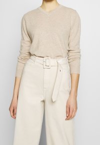 Beige kašmírový svetr s výstřihem do V, doplněný světle béžovými kalhotami s vysokým pasem a širokými nohavicemi. Kalhoty mají odpovídající pásek se čtvercovou sponou.