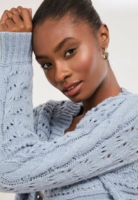 Svetlomodrý pletený kardigán s káblovými a čipkovými vzormi, so žebrovanými manžetami a uvoľneným šálovým golierom. Nosené zlaté kruhové náušnice.