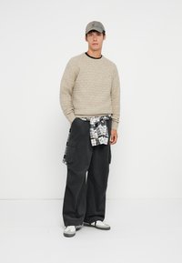 Beige texturovaný svetr, černé cargo kalhoty s kapsami, zavázaná kostkovaná košile, šedá kšiltovka a bílé tenisky. Jednoduchý, neformální outfit.