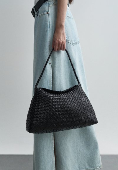 Mustakudos tullut käsilaukku, jossa on teksturoitu pinta, kaareva yläosa ja punottu kahva, jota pitää henkilö, joka on pukeutunut vaaleansiniseen farkkuhameeseen.