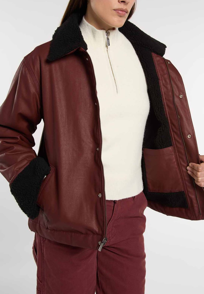 Giacca in pelle color borgogna con colletto e polsini in shearling nero; tasche laterali; chiusura frontale con zip; indossata sopra un maglione bianco con zip al collo.