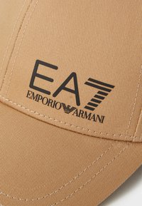 Khaki baseball cap with a textured fabric, featuring a black logo "EA7 EMPORIO ARMANI" on the front and contrasting white stitching.