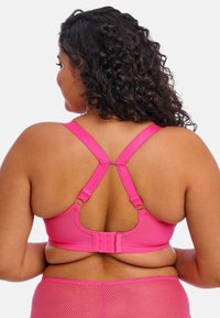 Světlý růžový podprsenka s nastavitelnými ramínky, s háčkovým a očkovým zapínáním na zadní straně a hladkým, pružným materiálem bez viditelných vzorů.