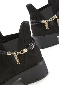 Črne gležnjarje iz semiša z elastičnimi stranskimi paneli, okrašene z pletenimi trakovi in zlatimi kovinskimi dodatki ter s čari v obliki ribje kosti.