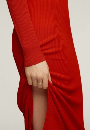 Mano con un anillo tirando de la abertura de un largo vestido rojo acanalado, mostrando parte de la pierna contra un fondo liso.