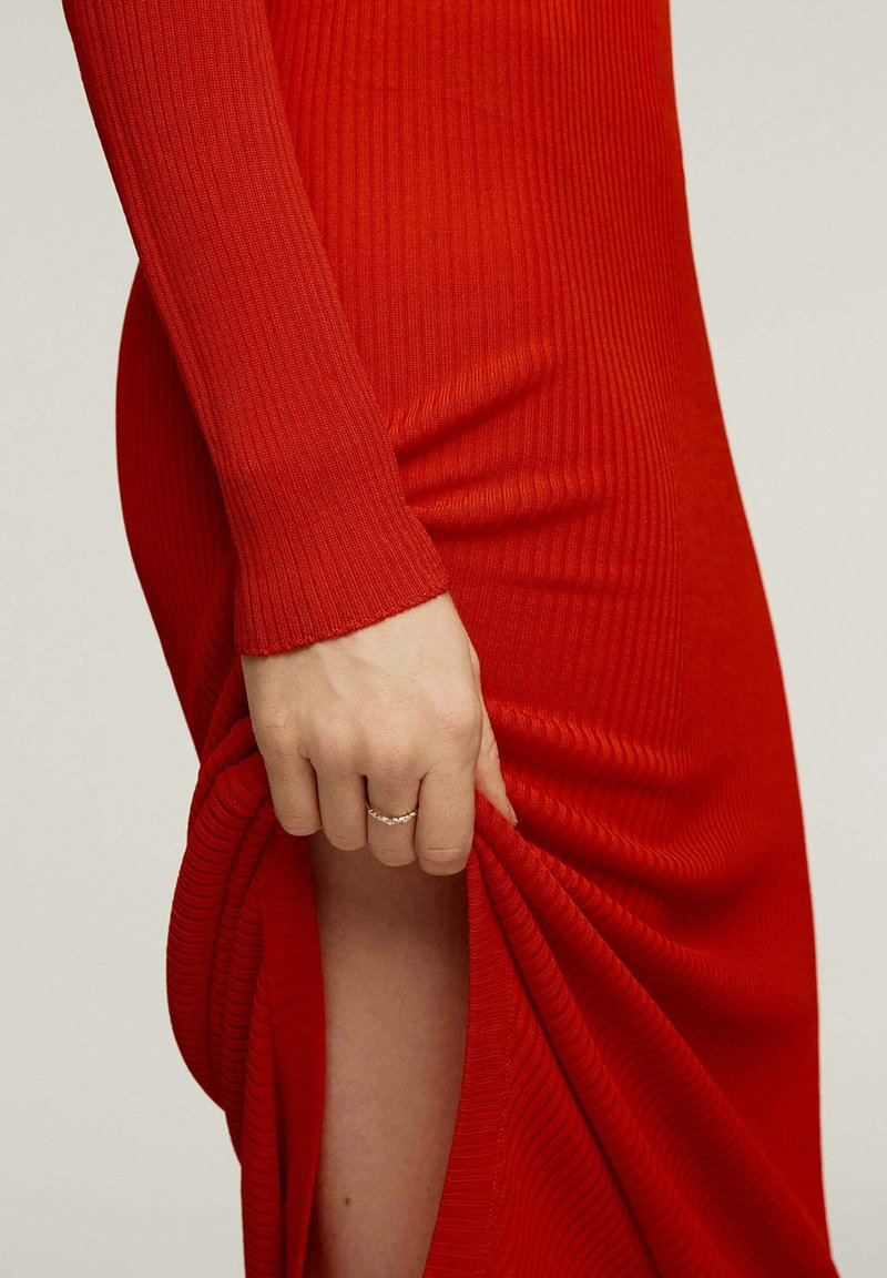 Mano con un anillo tirando de la abertura de un largo vestido rojo acanalado, mostrando parte de la pierna contra un fondo liso.