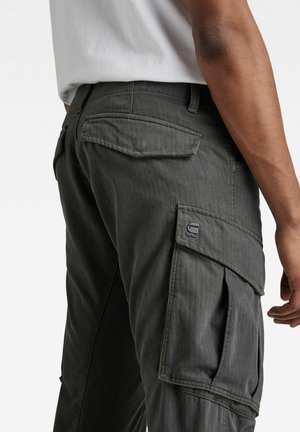 Zbliżenie osoby noszącej ciemnoszare spodnie typu cargo z klapkami na kieszeniach i małym logo na bocznej kieszeni, w połączeniu z białą koszulą.