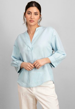 Blusa de manga larga azul clara con escote en V y hem con ligero redondeo. Está confeccionada en un material suave con una textura lisa y mínimos detalles de costura.