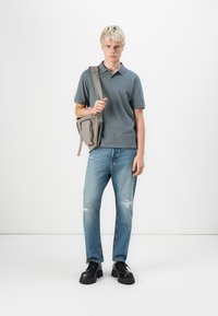 Grå polo shirt, blå slidte jeans og sorte sko med tykk sål. En grå crossbody taske hænger fra venstre skulder. Ensfarvet hvid baggrund.